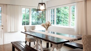 Un espace repas comprenant une table en bois, des chaises, un luminaire suspendu et de grandes fenêtres avec des rideaux.
