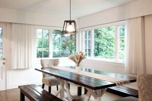 A dining area with a wooden table, chairs, a hanging light fixture, and large windows with curtains.