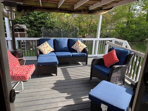 A covered outdoor deck with wicker furniture and cushions, a wooden fence, and a view of trees and a house in the background.