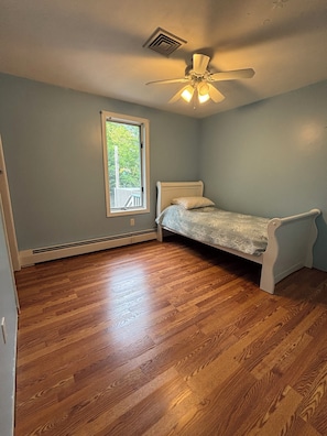 A bedroom with a single bed, a ceiling fan, a window with a view of greenery, and hardwood flooring.