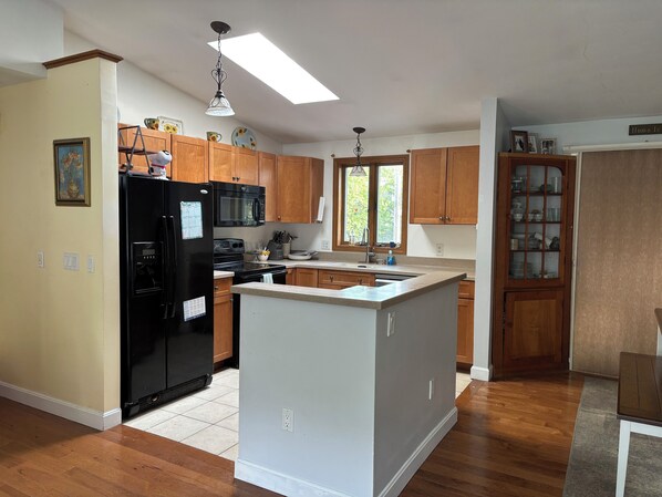 A kitchen with wooden cabinets, a black refrigerator, a microwave, and a window with a view of greenery.