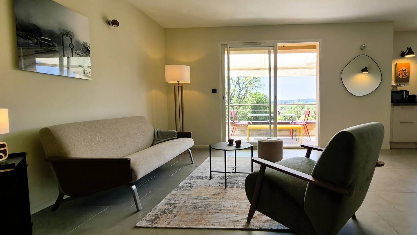 Ein Wohnzimmer mit Sofa, Sessel, kleinem runden Tisch, Stehlampe und Blick durch eine Glastür.