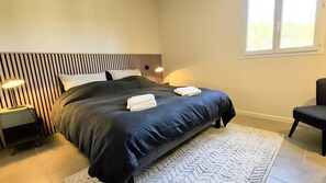 A modern bedroom with a striped headboard, a large bed with dark bedding, two bedside lamps, a window with a view, and a chair. - Pinarello Apartment. Sleeps 4, swimming pool, 500 m from the beach. (STE LUCIE DE PORTO VECCHIO)