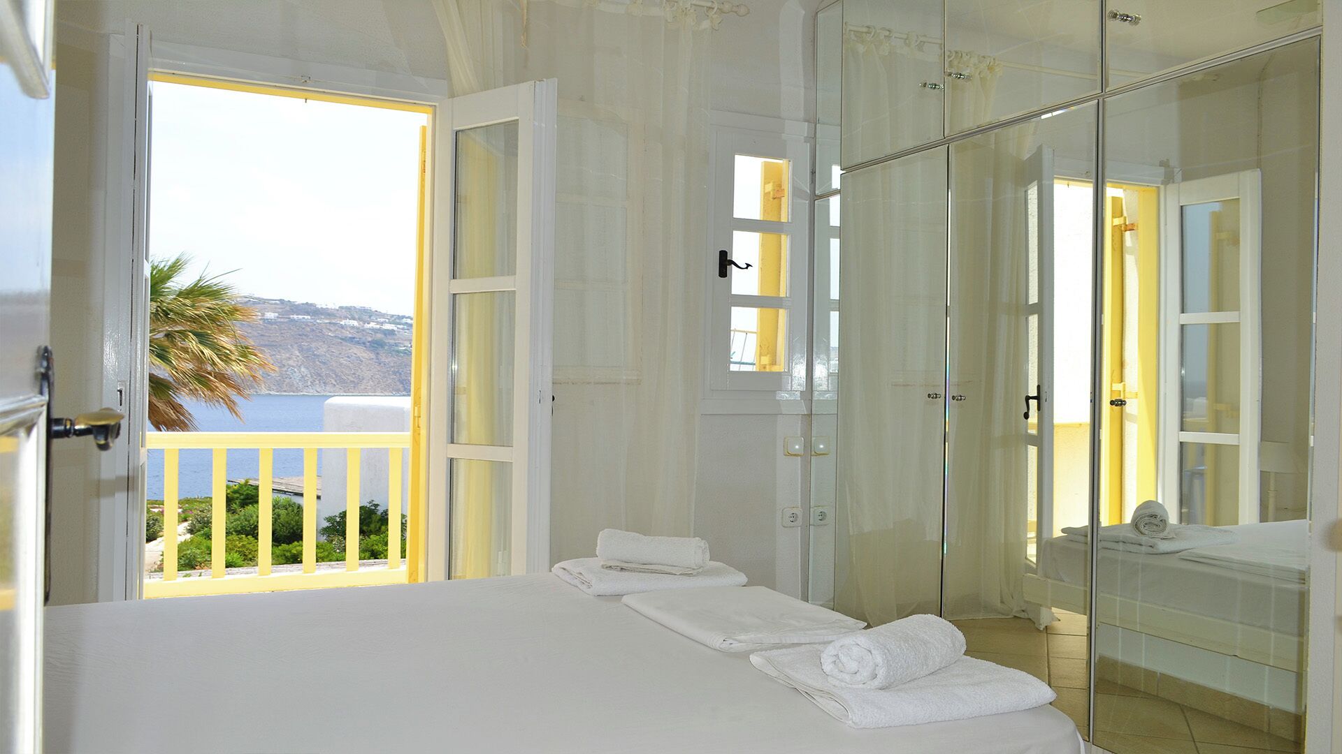 Quarto de hotel com varanda, cama com roupa de cama branca, chuveiro em cabine de vidro e vista para o mar.