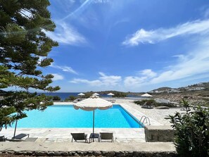 Ein Schwimmbecken mit Liegestühlen und Sonnenschirmen, umgeben von Steinmauern und Grünpflanzen, mit Blick auf das Meer und die Hügel im Hintergrund.