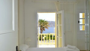 Ein Schlafzimmer mit Bett, Nachttisch, Lampe, einem Balkon mit Blick auf das Meer und eine Palme sowie einer Tür mit Fenster.