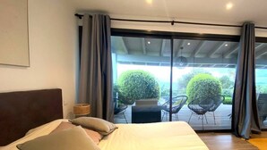 A bedroom with a large bed, a headboard, a bedside table with a lamp, and a view of an outdoor deck with chairs and greenery. - VILLA GIRELLES 5 Bed Room (PIETROSELLA)