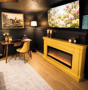 Une pièce avec un bureau en bois, une cheminée jaune, un téléviseur fixé au mur et un tableau encadré accroché au mur.