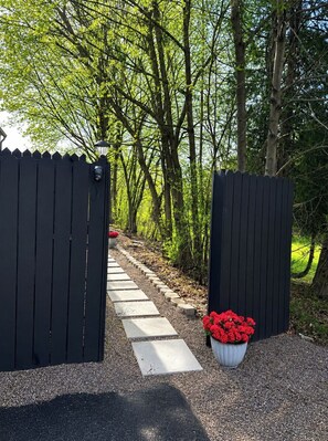 Een omheind pad dat leidt naar een bebost gebied, met een lantaarnpaal en rode bloemen.