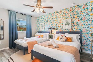 Habitación de hotel con dos camas, un ventilador de techo, una ventana con cortinas, una pared con un mosaico colorido y una mesita con una lámpara.