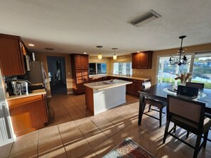 A kitchen with wooden cabinets, a central island, a dining area with chairs, and a view through a window. - Waterfront west coast home (Holiday)