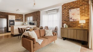 A cozy living room with a brown sofa, a dining area with a wooden table, and a kitchen with a brick wall. - North Street Panorama -stunning mountain and ocean views (Urunga)