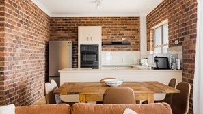 A compact kitchen with brick walls, a wooden dining table, and modern appliances. - North Street Panorama -stunning mountain and ocean views (Urunga)