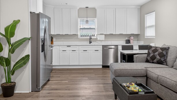Una cocina moderna con armarios blancos, un refrigerador de acero inoxidable y una zona de comedor con un sofá gris y una bandeja de aperitivos.