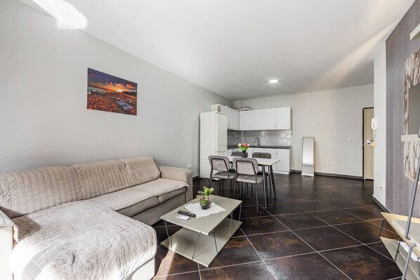 A modern living room with a sofa, a dining table with chairs, a kitchen area with cabinets, and a painting on the wall. - Sunset Apartment | 1BR Escape by PikHost (Durrës)