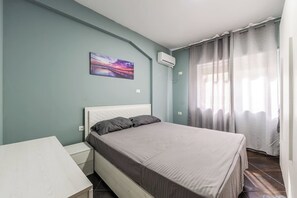 A bedroom with a bed, a bedside table, a wall-mounted air conditioner, and a window with curtains. - Sunset Apartment | 1BR Escape by PikHost (Durrës)