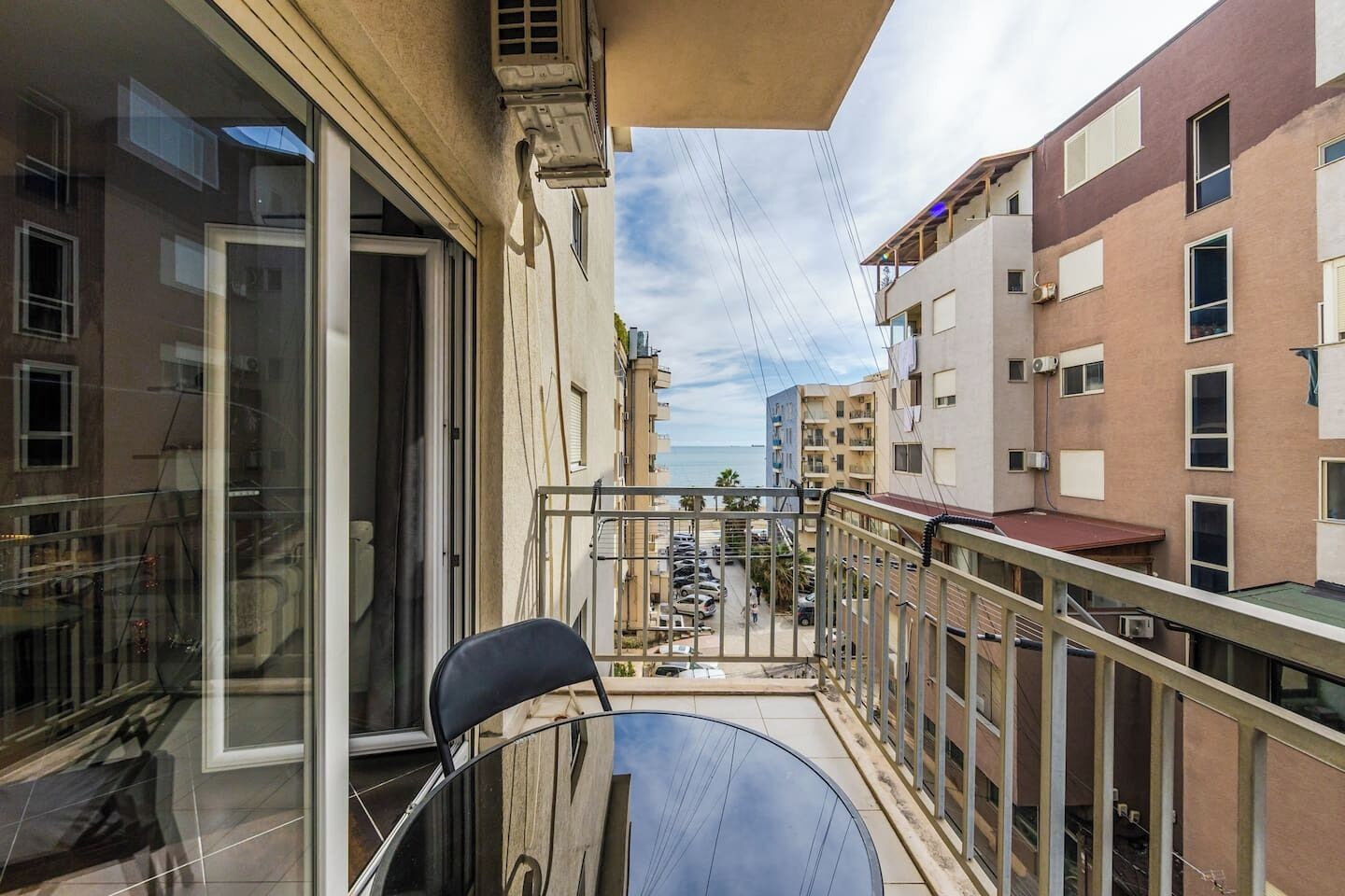 A balcony with a chair, a glass door, and a view of a street with parked cars and buildings.