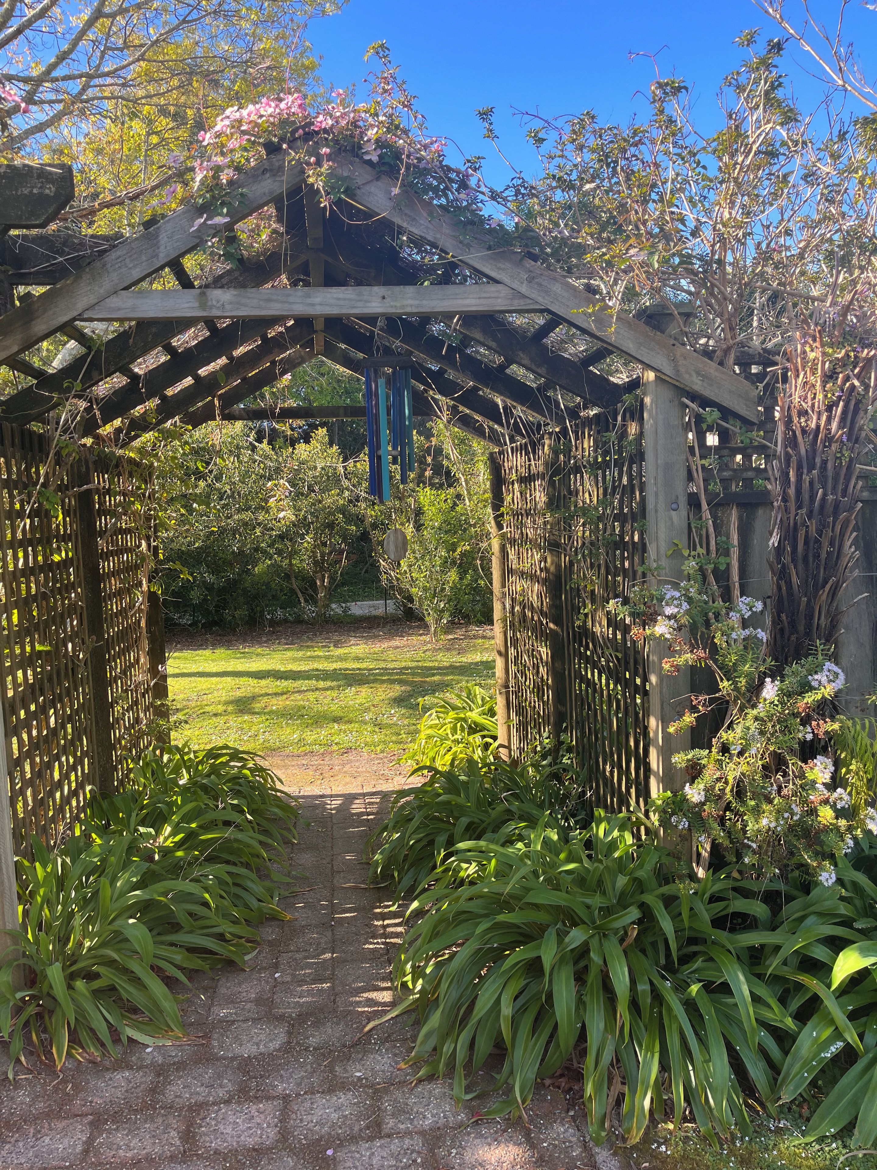 Een houten pergola met een lattenstructuur, leidend naar een tuin vol planten en een heldere lucht.