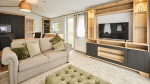 A modern living room with a sofa, ottoman, TV, wooden cabinets, and a kitchen area. - Host Stay Rose Lodge (York)