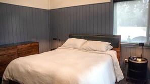 A bedroom with a bed, a nightstand, a lamp, and a window with a view. - Superior Queen Two-Bedroom Cottage - no pets (Bronson)