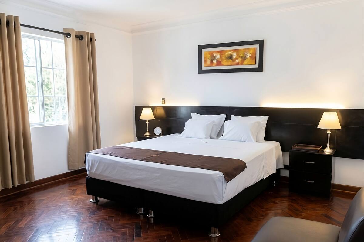 A hotel room with a large bed, two bedside tables with lamps, a framed artwork on the wall, and a view of greenery outside.