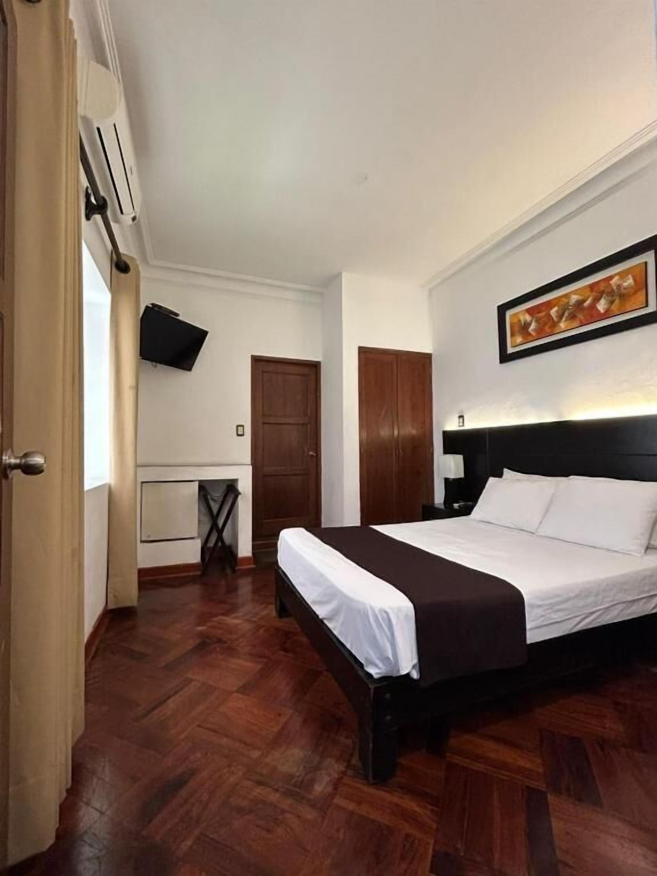 A hotel room with a bed, a desk, a chair, a TV mounted on the wall, and a door.