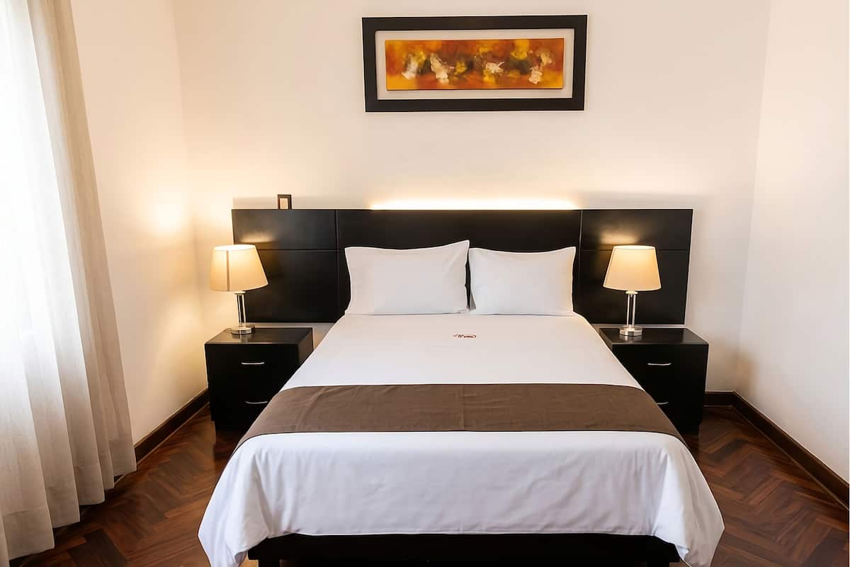 A hotel room with a large bed, two bedside tables with lamps, a headboard, a framed artwork on the wall, and a wooden floor.