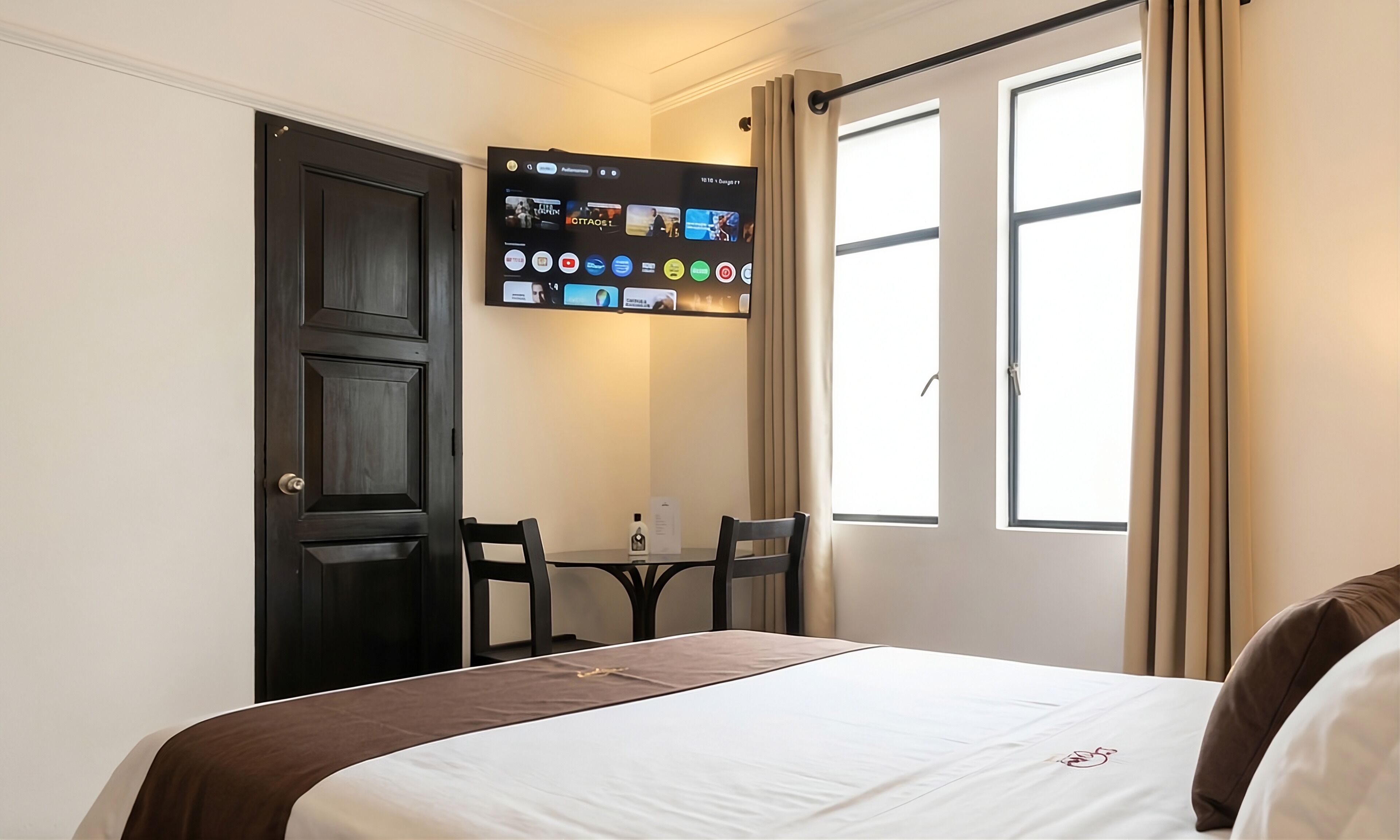 A hotel room with a bed, a desk with a computer, a chair, and a window with curtains.