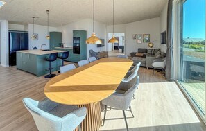 Eine moderne Küche mit Kochinsel, ein Essbereich mit großem Holztisch und ein Wohnzimmer mit Sofa und Fernseher.