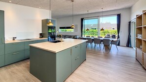 Cocina moderna con isla central, zona de comedor con sillas y ventanas amplias que ofrecen vistas al verdor.