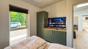 Un dormitorio con cama, televisor sobre un mueble, ventana que ofrece vistas a un coche y follaje, y puerta que da a otra habitación.