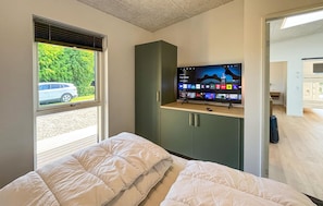 Ein Schlafzimmer mit einem Bett, einem Fernseher auf einer Kommode, einem Fenster mit Blick auf ein Auto und Grünflächen sowie einer Tür zu einem anderen Raum.