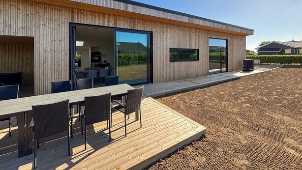 Un edificio moderno con amplios ventanales, una terraza de madera con mobiliario exterior y una vista despejada del paisaje circundante.