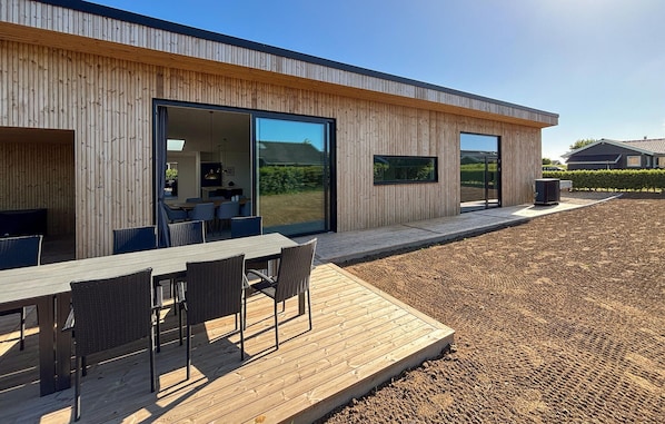 Ein modernes Gebäude mit großen Glasüren, einer Holzterrasse mit Gartenmöbeln und freiem Blick auf die umliegende Landschaft.