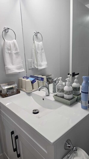 Une salle de bain avec un lavabo blanc, un miroir, deux porte-serviettes, un distributeur de savon et un flacon de gel hydroalcoolique.