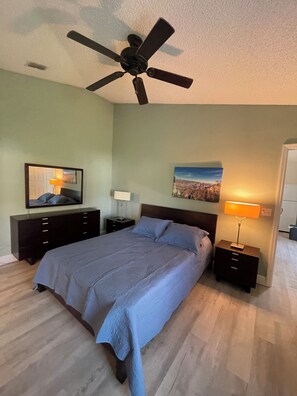 Ein Schlafzimmer mit Bett, Deckenventilator, Nachttisch mit Lampe, Kommode und einem Bild an der Wand.