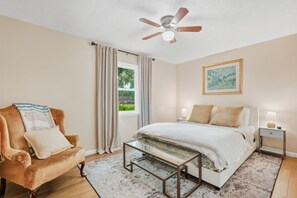 Ein Schlafzimmer mit einem Bett, einem Sessel, einem Deckenventilator, einem Fenster mit Vorhängen, einem Nachttisch und einem gerahmten Bild an der Wand.
