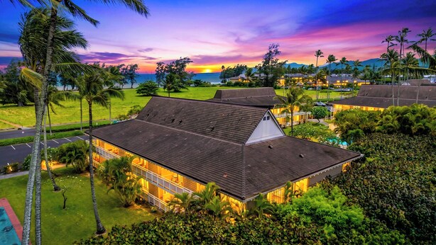 Ein Hotel mit geneigtem Dach, umgeben von üppiger Vegetation und Palmen, mit Blick auf das Meer bei Sonnenuntergang.