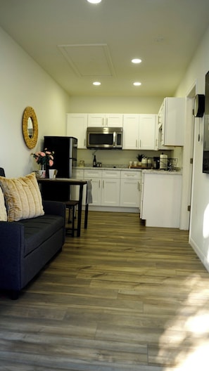 A compact living space with a dark sofa, a dining table, a kitchen area with white cabinets and a microwave, and a wooden floor.