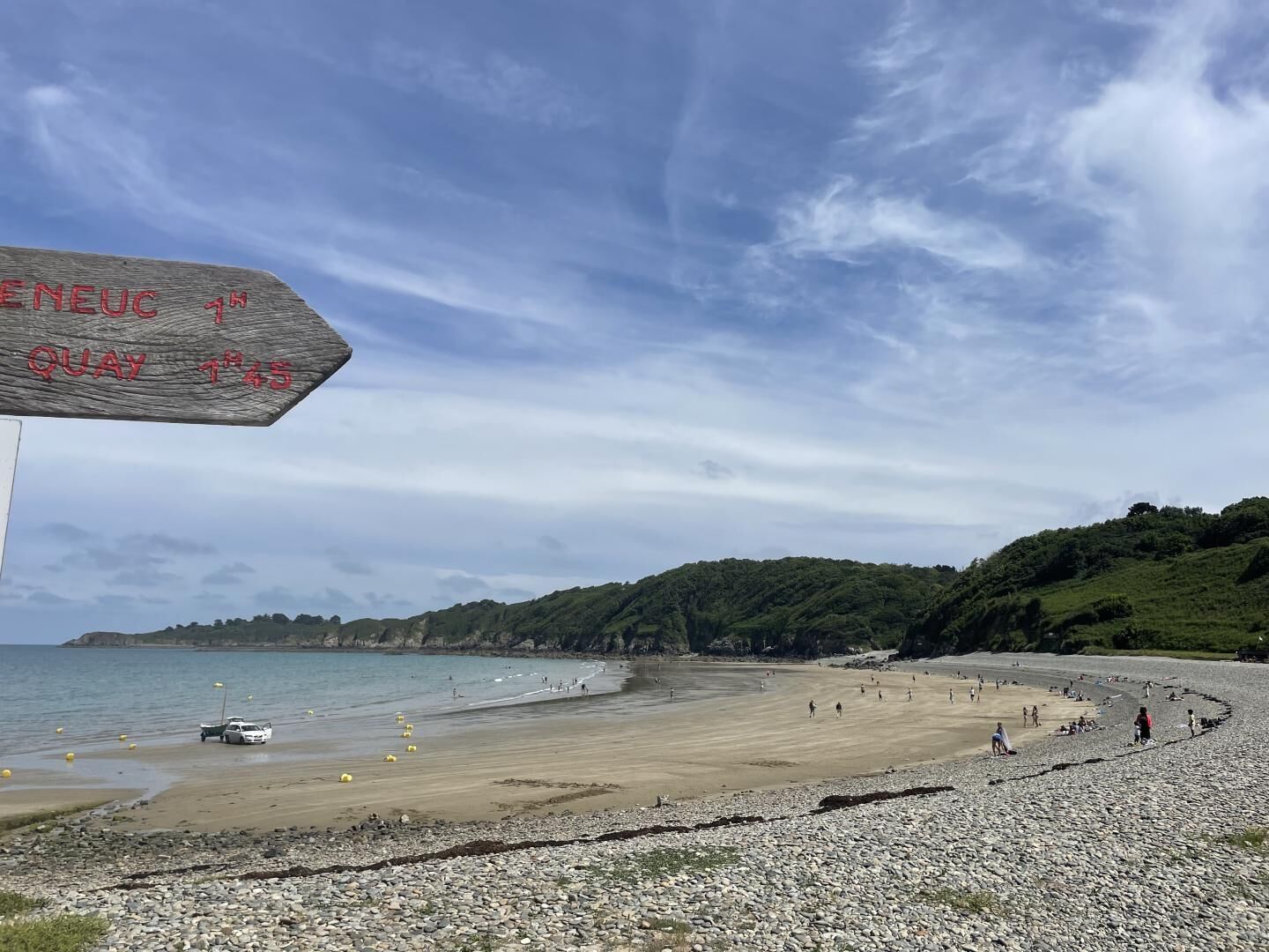 A beach with a wooden sign, pebbles, and people.