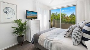 Ein Schlafzimmer mit Bett, Fernseher, Deckenventilator, Topfpflanze und einem Balkon mit Blick auf Grünflächen.