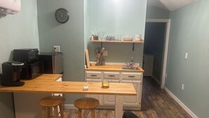 Eine kleine Küche mit einem Holzbarhocker, einer Mikrowelle, einer Kaffeemaschine und einem Mülleimer.