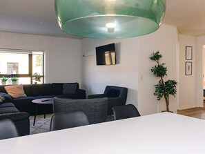 Ein modernes Wohnzimmer mit einer Couch, Sesseln, einem Tisch, einem an der Wand montierten Fernseher und einem großen Fenster mit Pflanzen.
