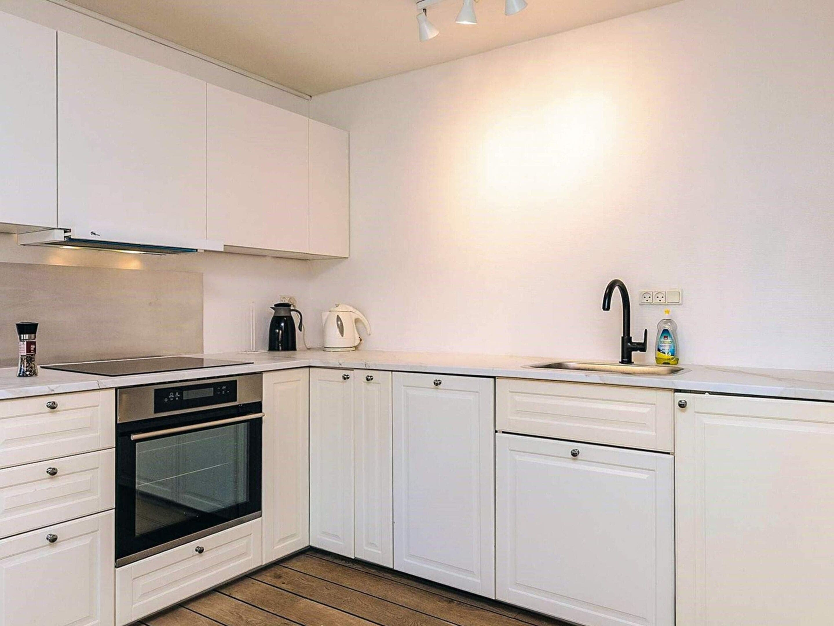 Una cocina moderna con muebles blancos, horno empotrado, lavamanos con grifo negro y hervidor en la encimera.