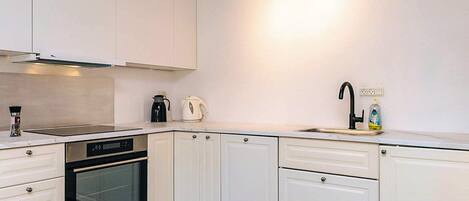 Una cocina moderna con muebles blancos, horno empotrado, lavamanos con grifo negro y hervidor en la encimera.