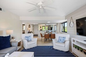 Ein modernes Wohnzimmer mit Deckenventilator, weißen Sofas mit gemusterten Kissen, einem Essbereich und einem an der Wand befestigten Fernseher.
