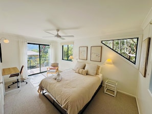 A bedroom with a bed, a desk, a chair, a ceiling fan, and large windows.