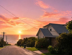 A residential house with a driveway leading to the front door, surrounded by greenery and a clear view of the ocean at sunset.