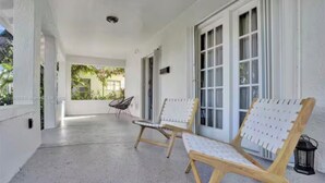 A covered porch with two wooden chairs and a table, white walls, and large windows.