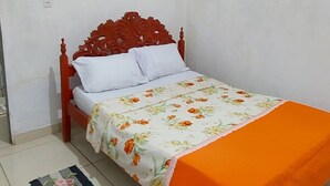 Una cama individual con una colcha estampada y un plaid naranja, cabecera de madera y una alfombra a rayas sobre suelo de baldosas.